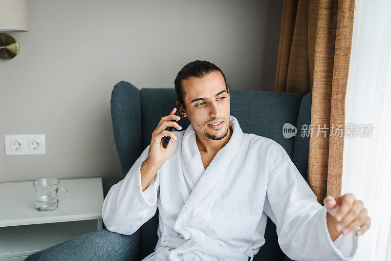 年轻有吸引力的白人男子/商人穿着浴袍坐在椅子上，谈论他的智能手机。